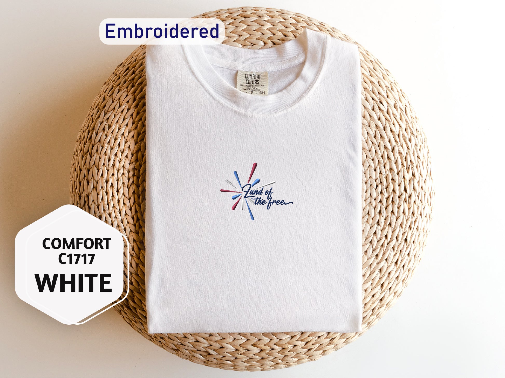 a white t-shirt with a colorful design, laid out on a woven basket or mat. The t-shirt has a label on the collar, and there is a hexagonal sticker next to it with text.