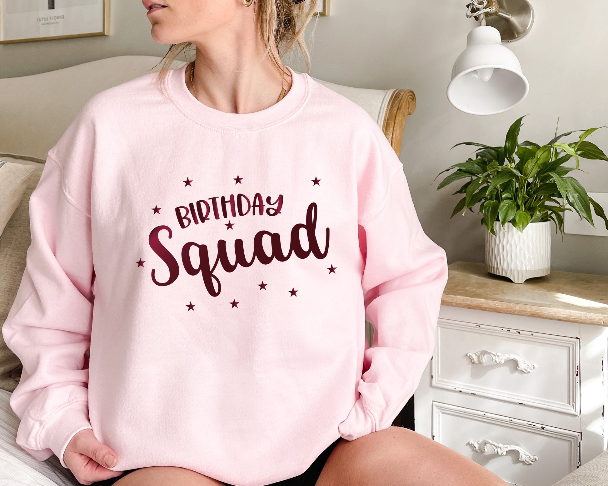 A woman wearing a pink sweatshirt with the text "Birthday Squad" printed on it, sitting on a bed in a bedroom setting.
