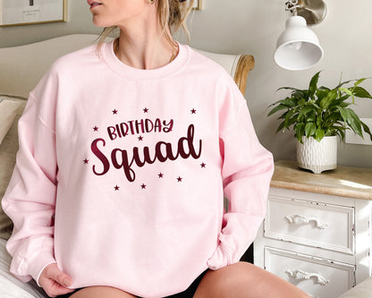 A woman wearing a pink sweatshirt with the text "Birthday Squad" printed on it, sitting on a bed in a bedroom setting.
