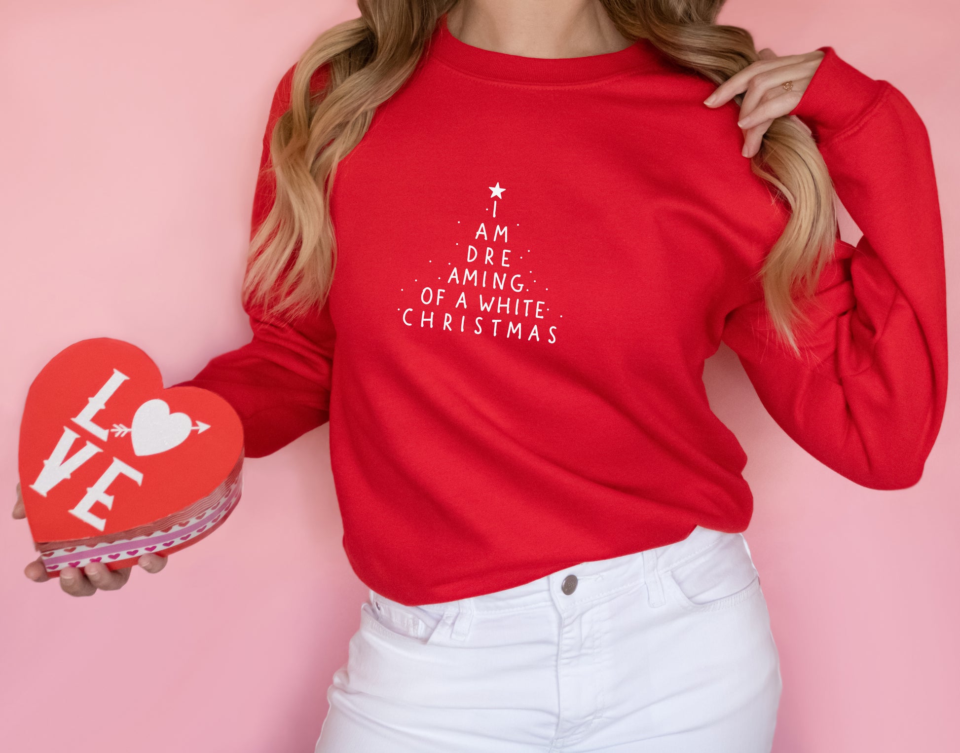 A woman wearing a red sweater with white text on it is holding a red heart-shaped box with the word "LOVE" written on it.