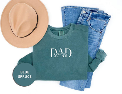 A casual outfit consisting of a green sweatshirt with the text "DAD" printed on it, a pair of blue jeans, and a tan hat. The items are arranged on a white background, with a blue circle labeled "BLUE SPRUCE" placed next to the outfit.