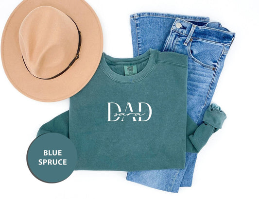A casual outfit consisting of a green sweatshirt with the text "DAD" printed on it, a pair of blue jeans, and a tan hat. The items are arranged on a white background, with a blue circle labeled "BLUE SPRUCE" placed next to the outfit.