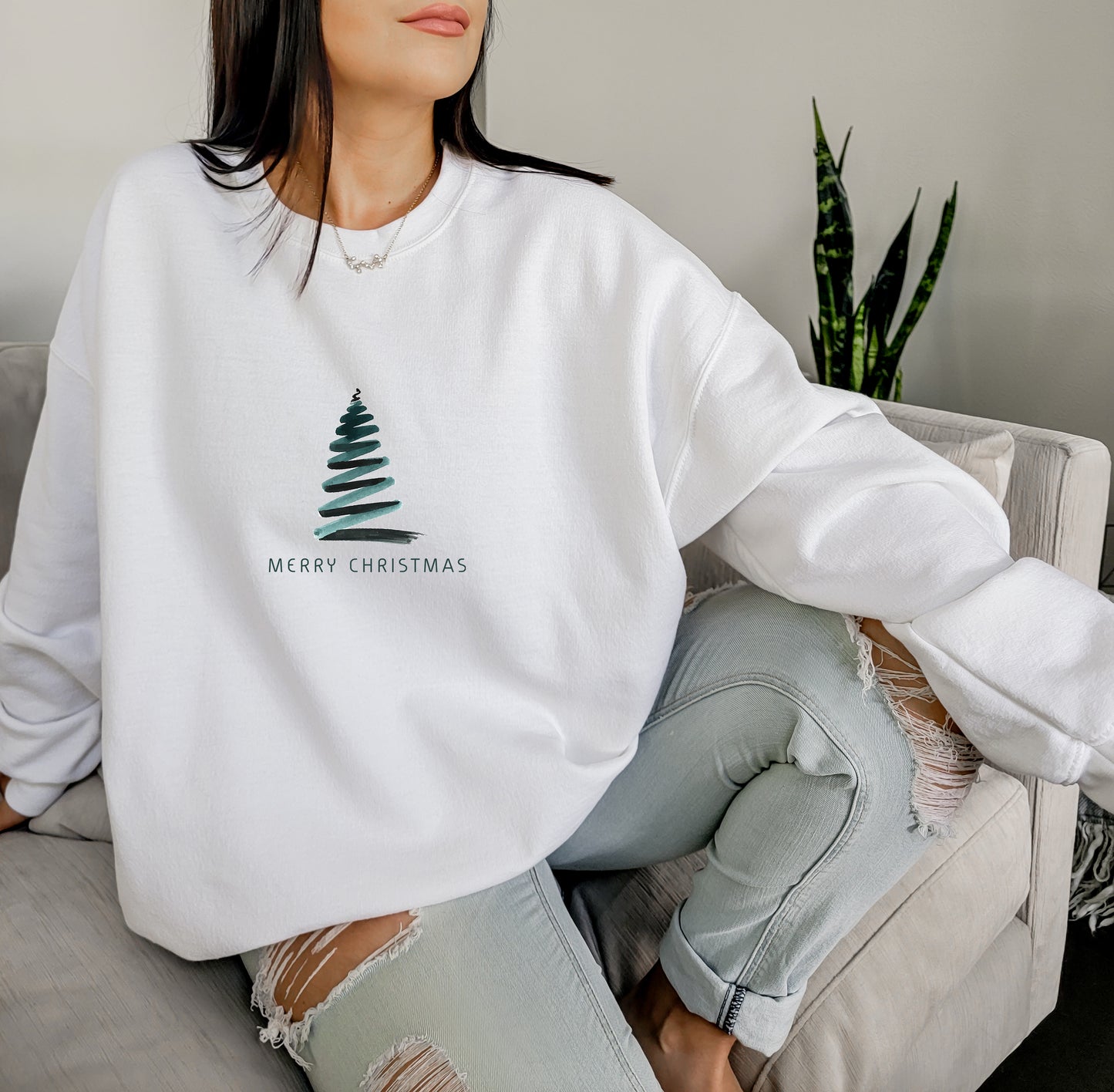 A woman wearing a white sweater with a Christmas tree design and the text "Merry Christmas" is sitting on a couch.