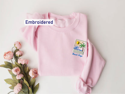 A pink sweater with a beach-themed embroidered patch is displayed on a white background, accompanied by a bouquet of pink flowers.