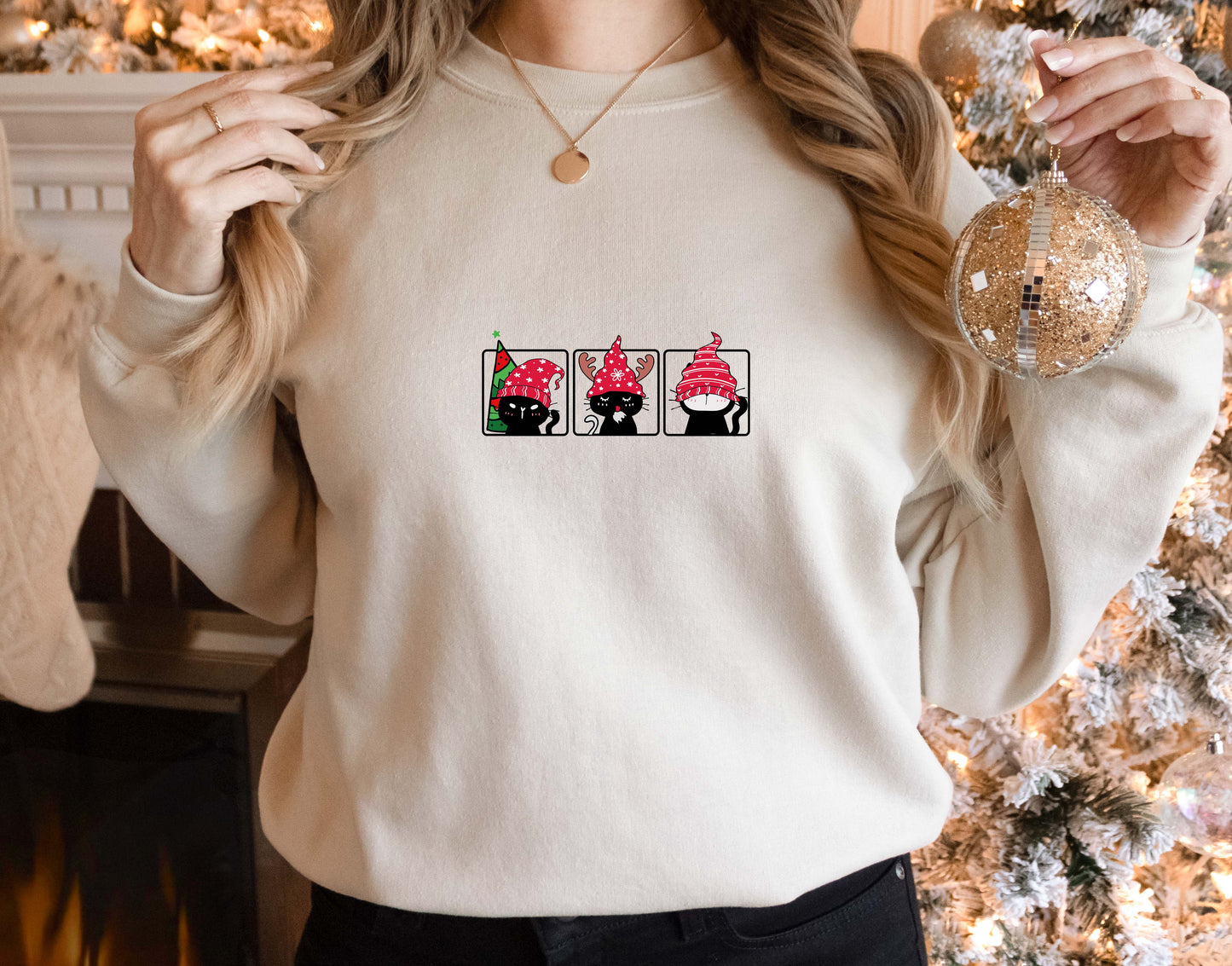 A woman wearing a white sweater with a graphic design of three cats wearing red hats. She is holding a Christmas ornament in her right hand.
