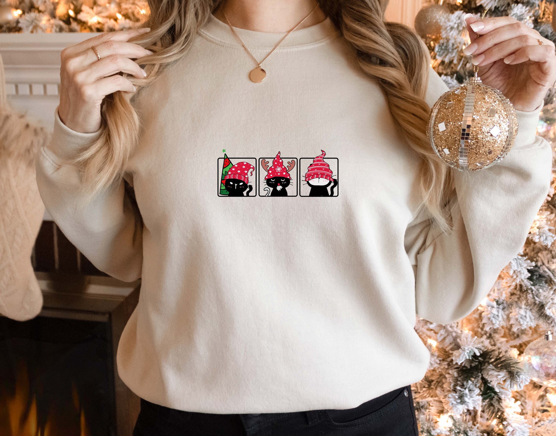 A woman wearing a white sweater with a graphic design of three cats wearing red hats. She is holding a Christmas ornament in her right hand.