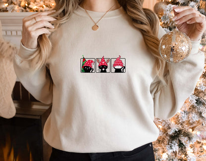 A woman wearing a white sweater with a graphic design of three cats wearing red hats. She is holding a Christmas ornament in her right hand.