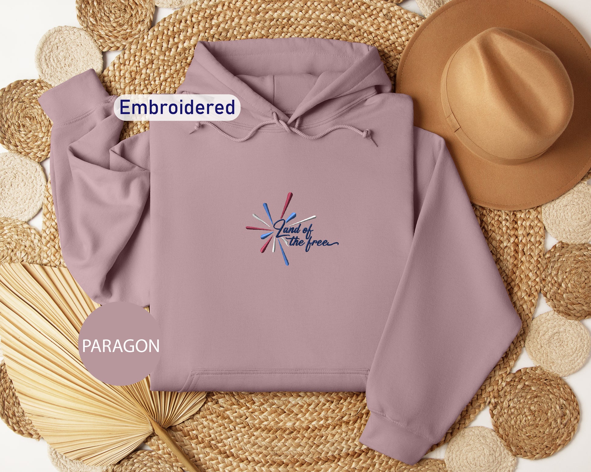 a purple hoodie with a patriotic design, a brown hat, and a woven straw hat, all arranged on a woven mat.