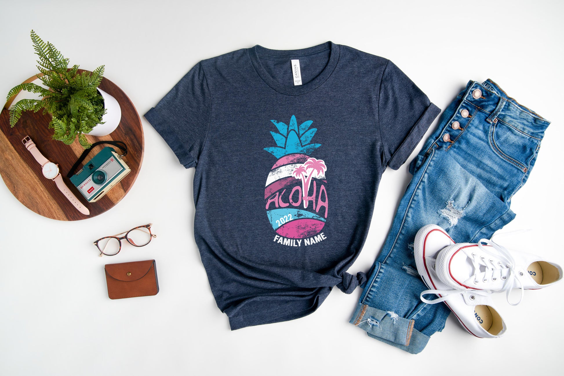 a casual outfit consisting of a blue t-shirt with a graphic design, a pair of jeans, a pair of white sneakers, a pair of glasses, a brown wallet, and a small plant.
