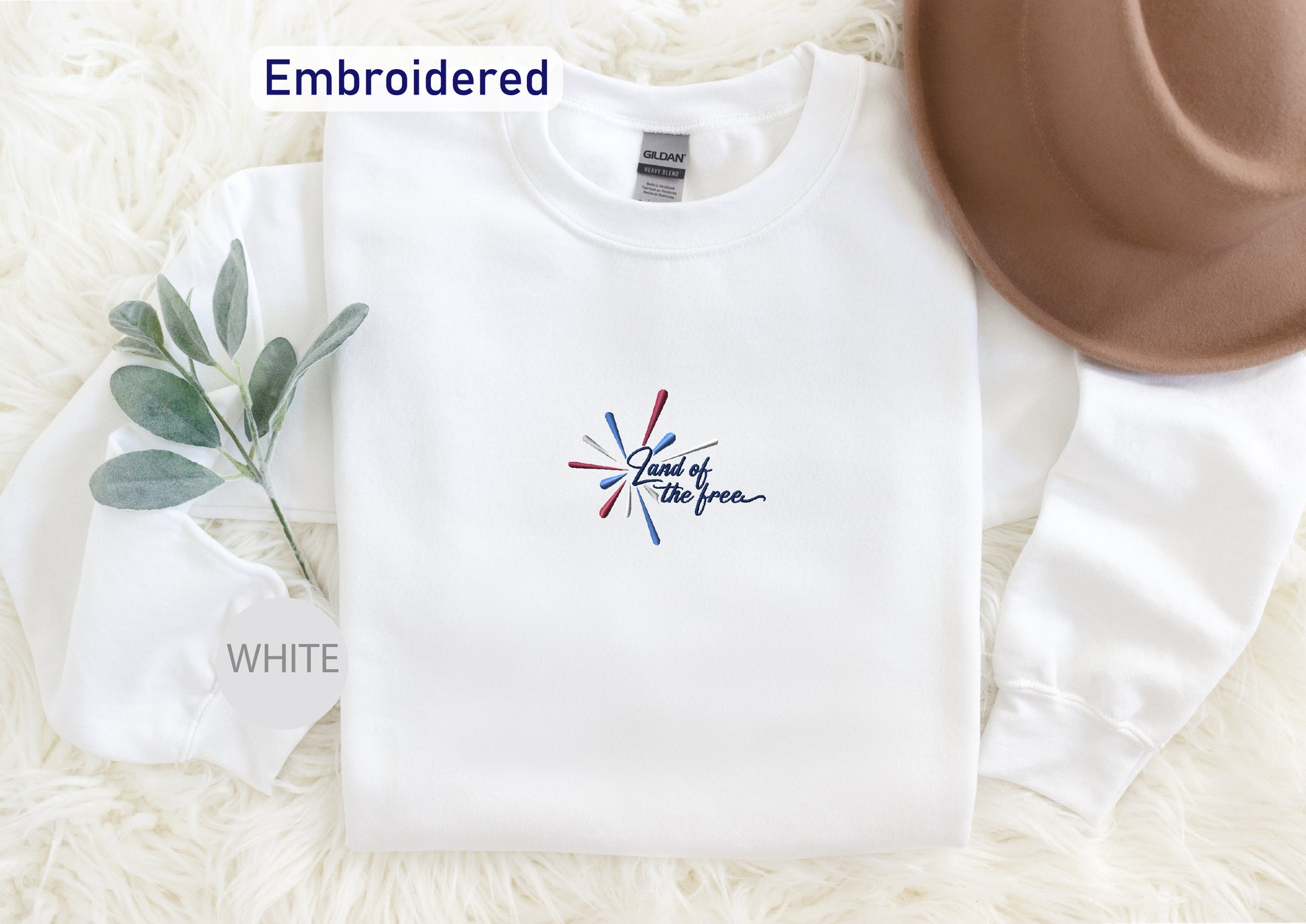 A white long-sleeved shirt with a colorful embroidered design, a brown hat, and a white sock are arranged on a white fur-covered surface.