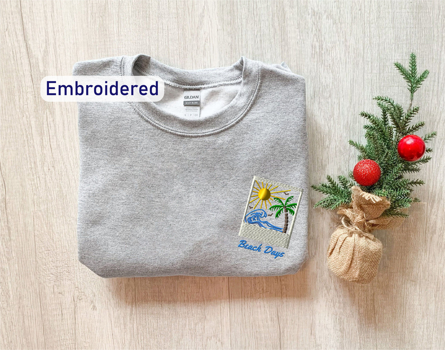 a gray sweatshirt with a beach-themed embroidered patch on the front, placed next to a small Christmas tree with a red ornament.