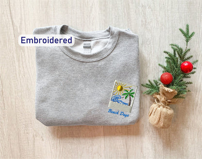 a gray sweatshirt with a beach-themed embroidered patch on the front, placed next to a small Christmas tree with a red ornament.