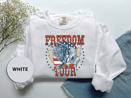 a white long-sleeved shirt with a graphic design that says "FREEDOM FOUR" and an eagle with an American flag design. The shirt is laid out on a white surface, with a pair of denim jeans visible in the background.