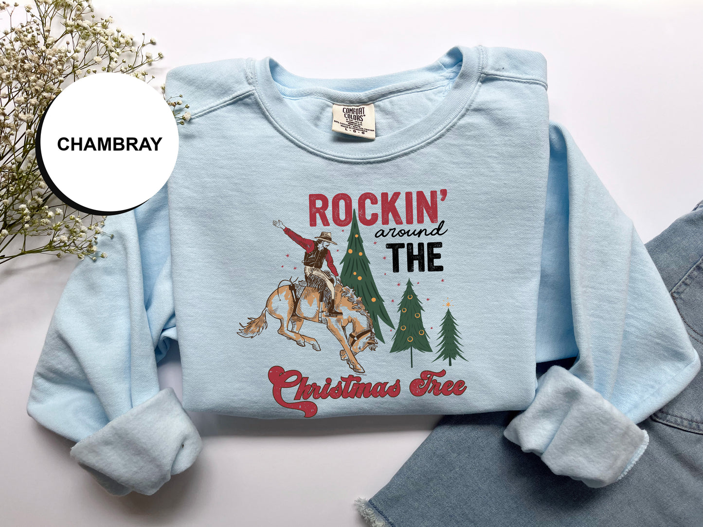 A light blue long-sleeved shirt with a graphic design depicting a cowboy on a horse, surrounded by Christmas trees, and the text "ROCKIN' around THE CHRISTMAS Tree".