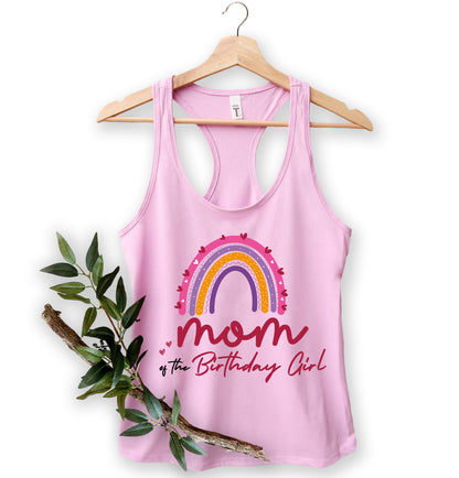 A pink tank top with a rainbow graphic and the text "Mom of the Birthday Girl" printed on it, hanging on a wooden hanger.