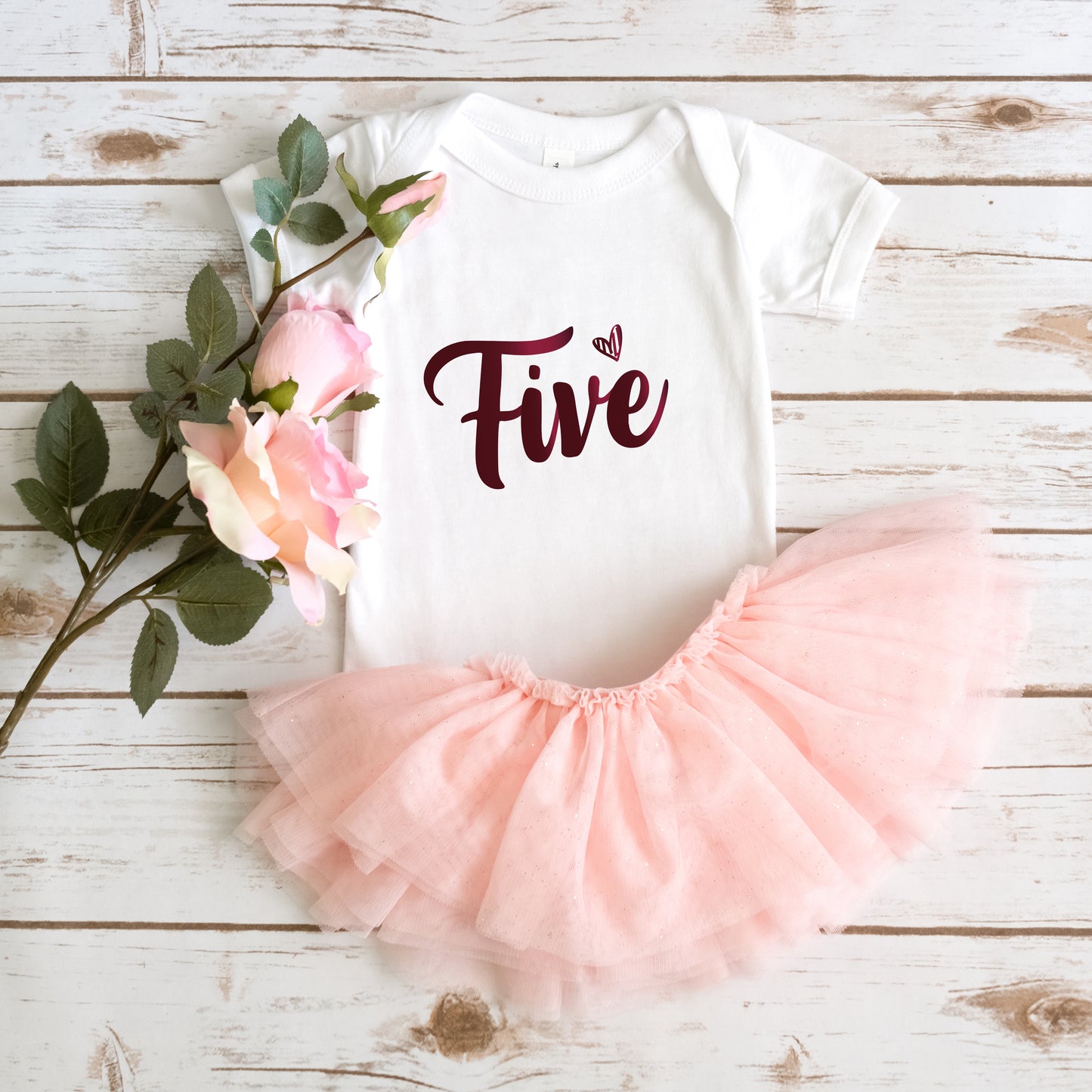 A white onesie with the number "Five" printed on it, a pink tutu skirt, and a bouquet of pink roses are arranged on a wooden surface.