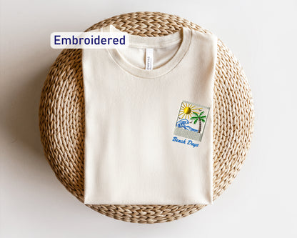 A white t-shirt with a beach-themed embroidered design is displayed on a woven basket.