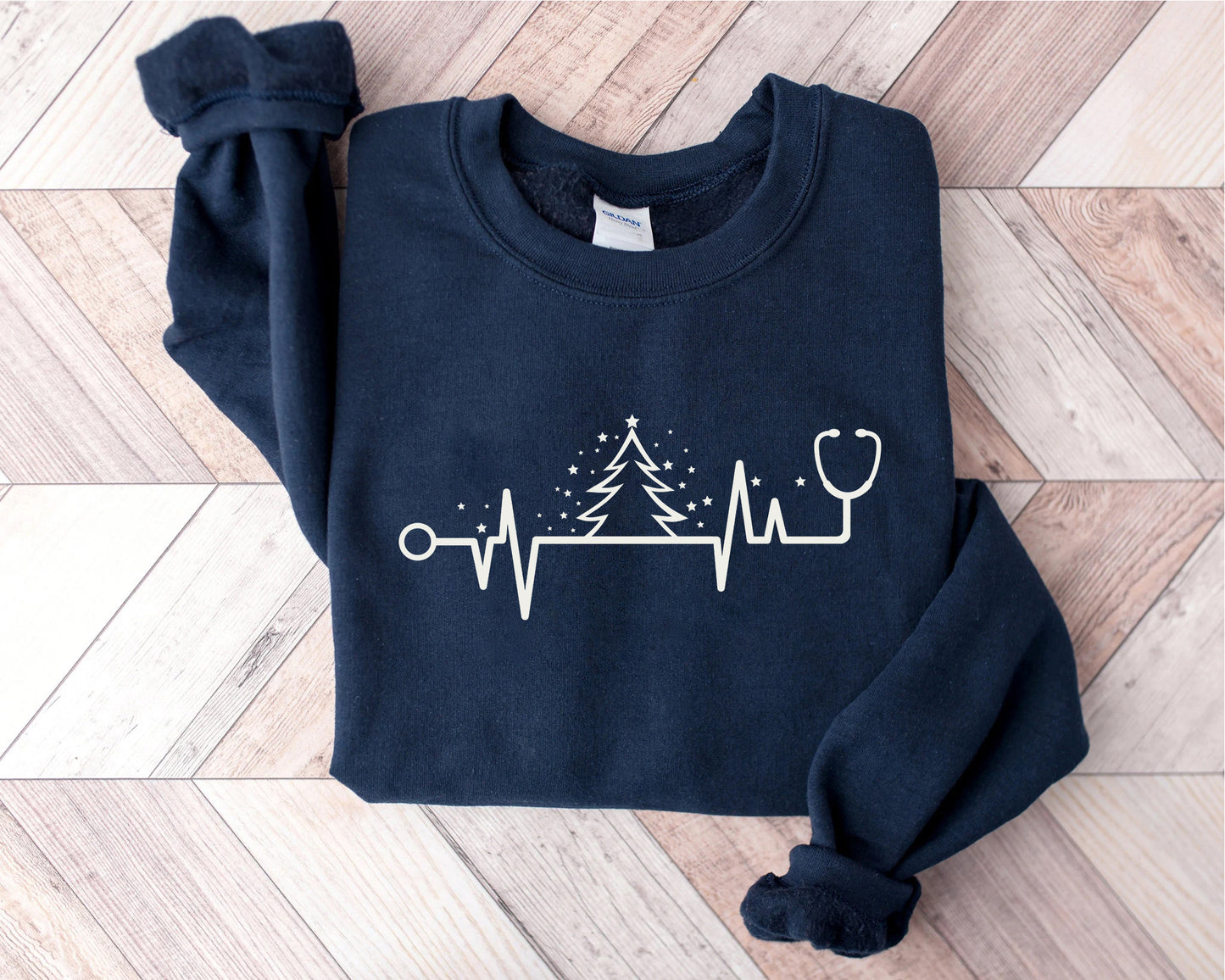 A navy blue sweatshirt with a white graphic design on it, depicting a Christmas tree and a stethoscope, is laid out on a wooden surface.
