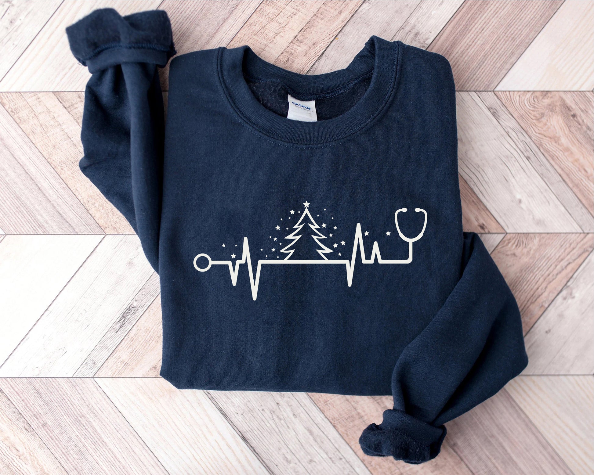 A navy blue sweatshirt with a white graphic design on it, depicting a Christmas tree and a stethoscope, is laid out on a wooden surface.