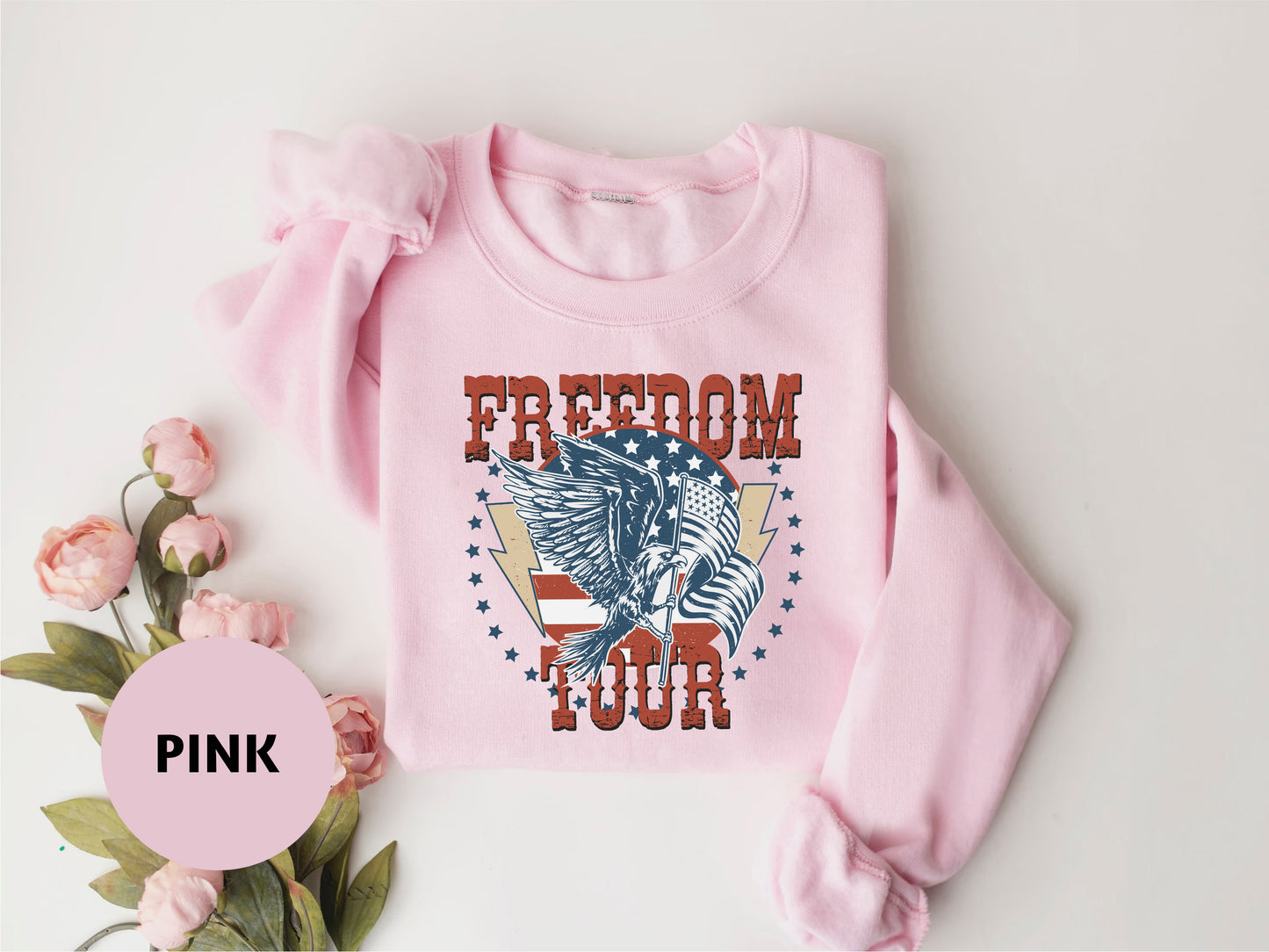 A pink sweatshirt with a graphic design featuring an eagle and the text "FREEDOM TOUR" is displayed on a white background.