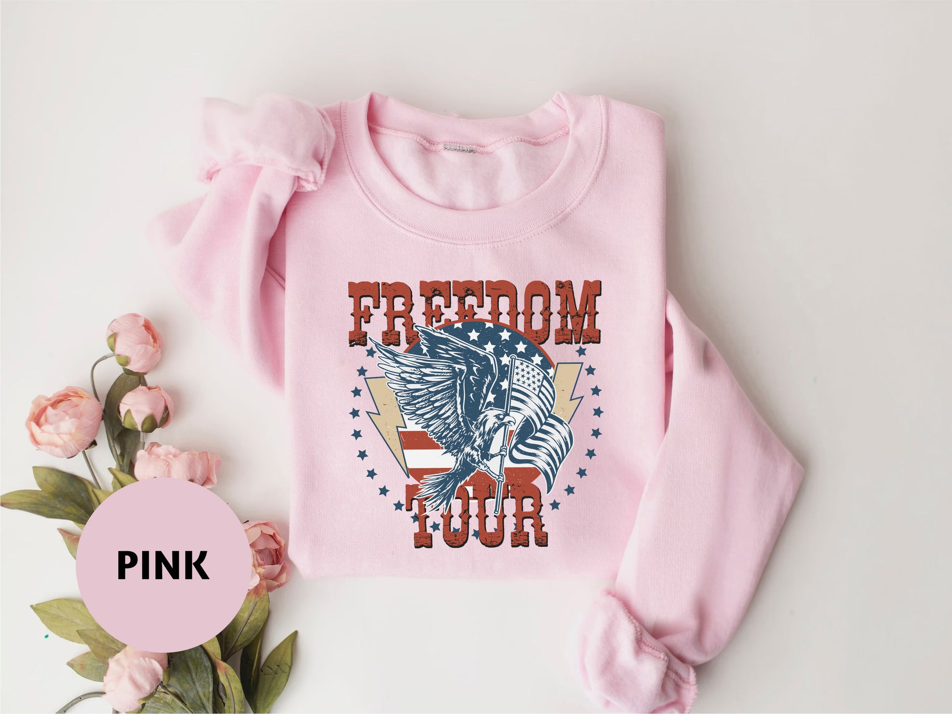 A pink sweatshirt with a graphic design featuring an eagle and the text "FREEDOM TOUR" is displayed on a white background.