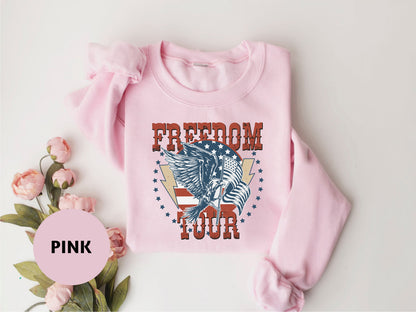 A pink sweatshirt with a graphic design featuring an eagle and the text "FREEDOM TOUR" is displayed on a white background.