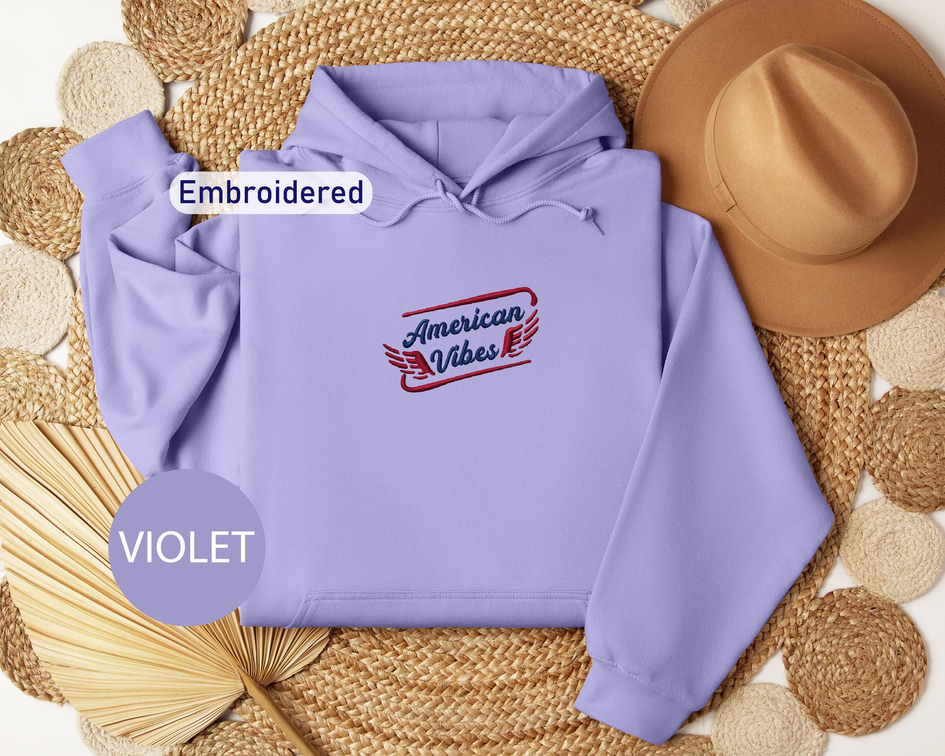 a purple hoodie with the text "American Vibes" embroidered on it, along with a brown hat and a straw hat placed on a woven mat.