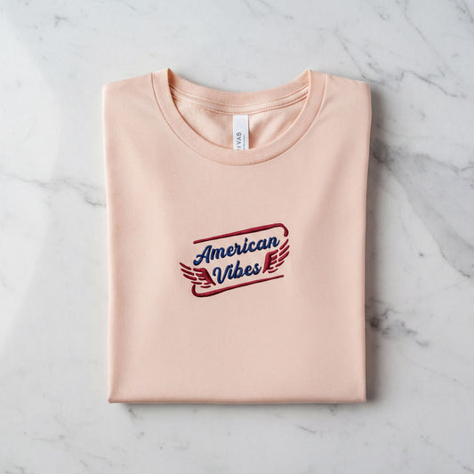 A pink t-shirt with the text "American Vibes" printed on it, laid out on a marble surface.