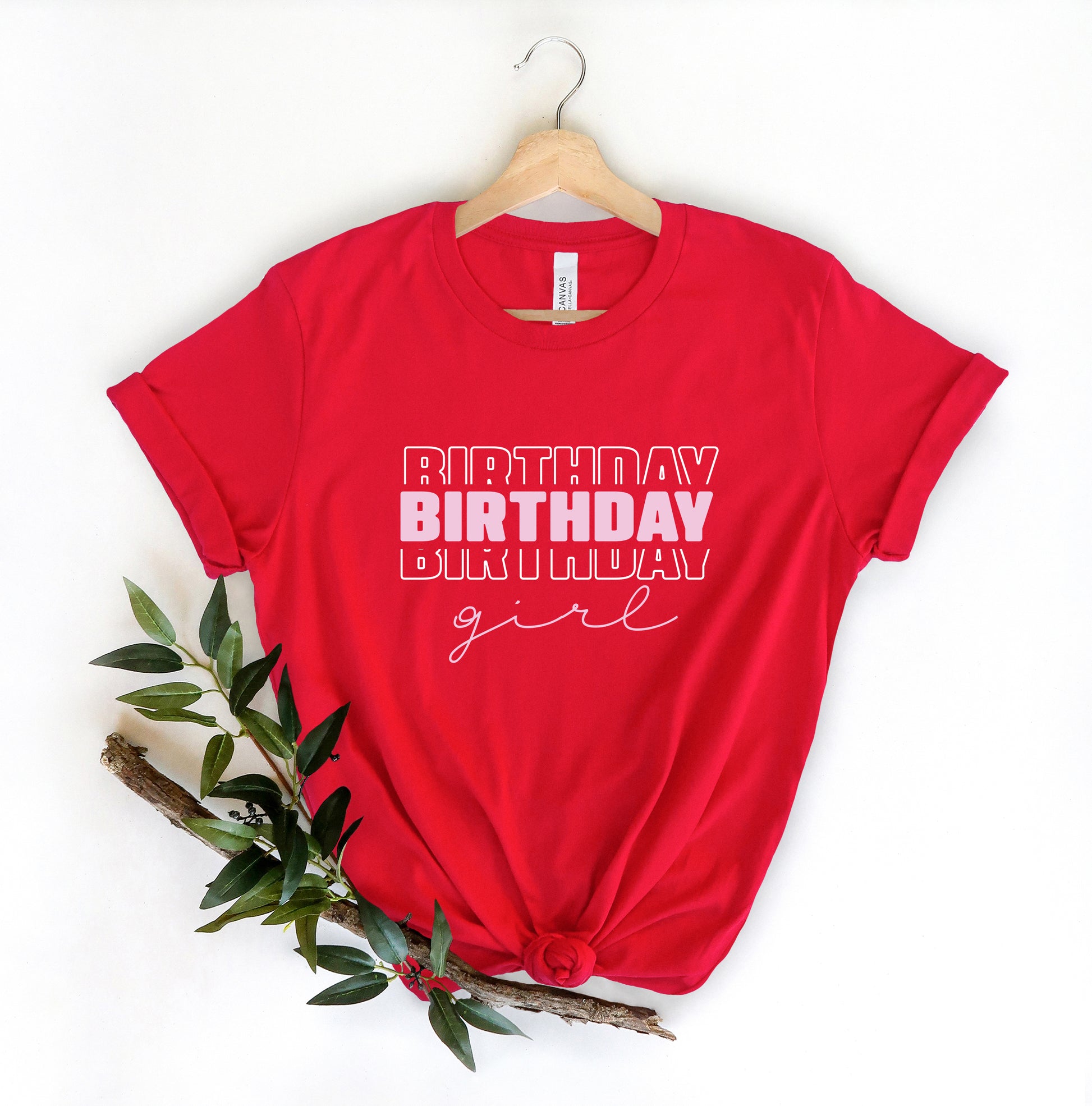 A red t-shirt with the text "BIRTHDAY GIRL" printed on it, along with a wooden hanger. The t-shirt is displayed against a white background, and there is a branch of green leaves and a small red flower nearby.