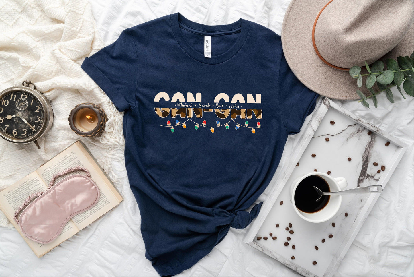 a casual arrangement of items, including a navy blue t-shirt with a graphic design, a hat, a book, a cup of coffee, and a pair of sunglasses.