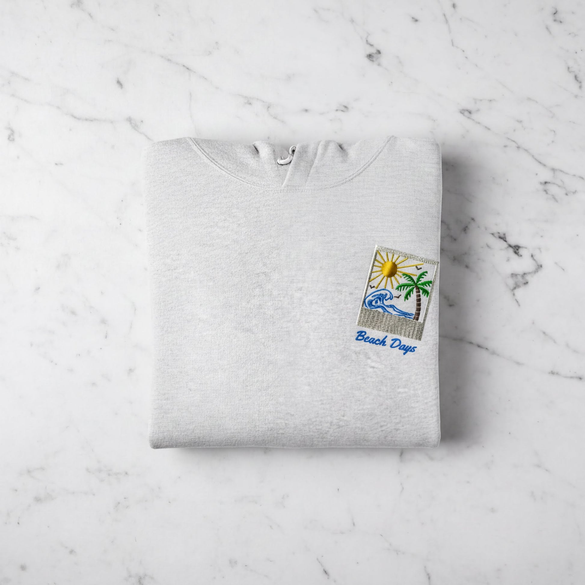 A white hoodie with a beach-themed design and the text "Beach Days" printed on it, laid out on a marble surface.