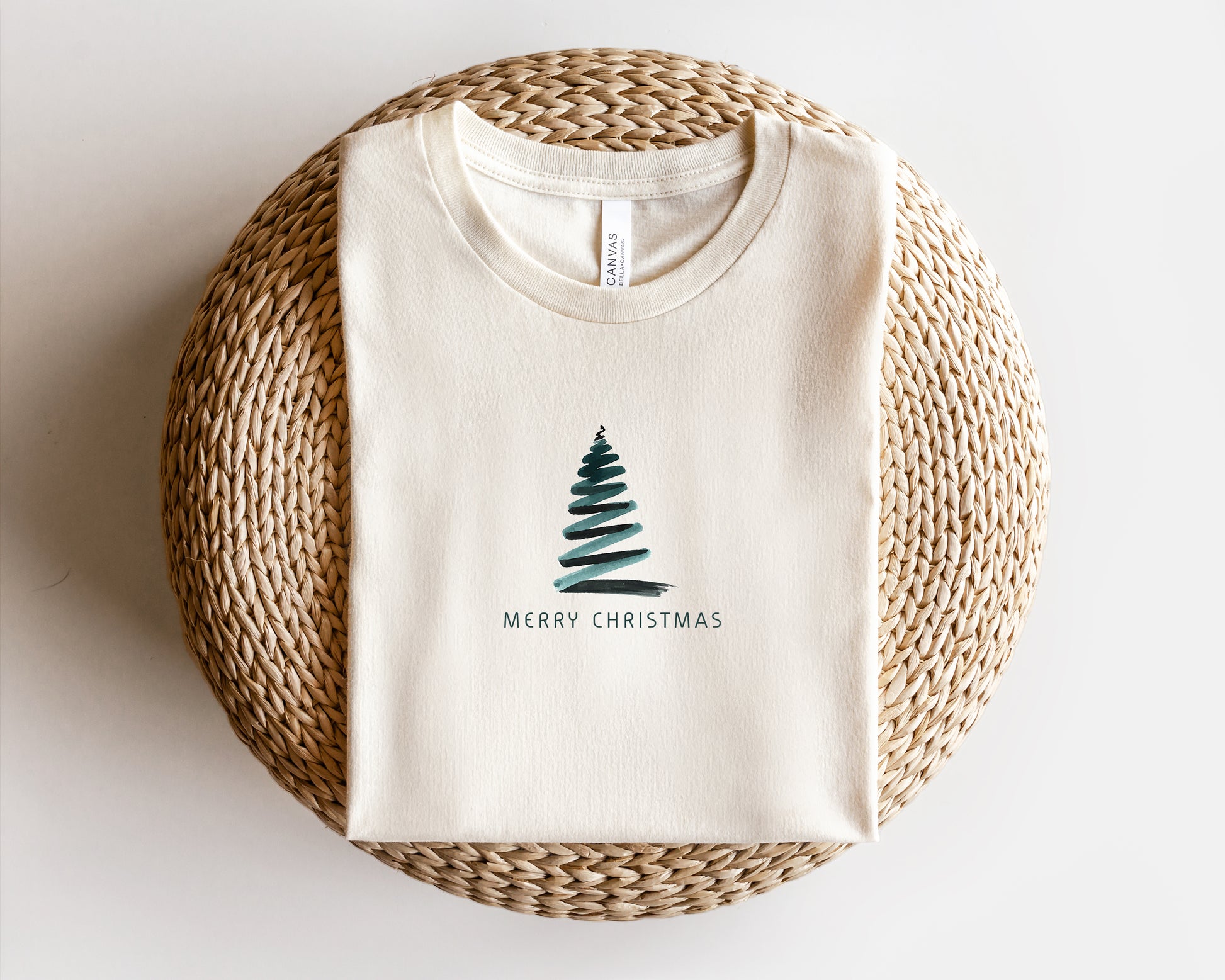 A white t-shirt with a Christmas tree graphic and the text "Merry Christmas" printed on it, laid out on a woven basket.