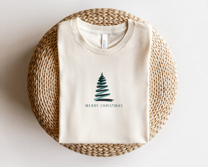 A white t-shirt with a Christmas tree graphic and the text "Merry Christmas" printed on it, laid out on a woven basket.