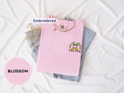 a pink t-shirt with a graphic design, a pair of denim jeans, and some dried flowers, all laid out on a white surface.