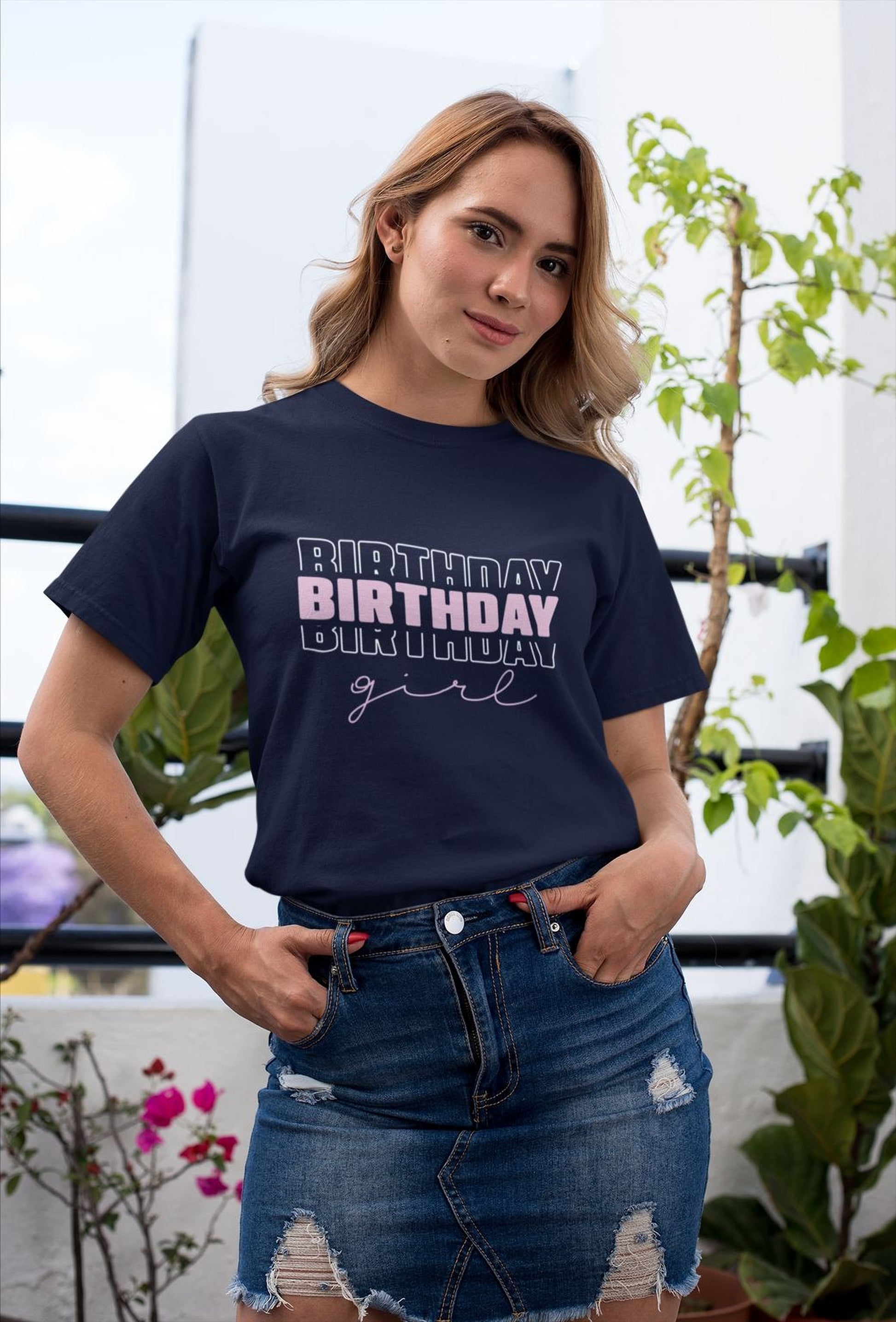 A young woman wearing a navy blue t-shirt with the text "BIRTHDAY GIRL" printed on it, standing in front of a white wall with plants.