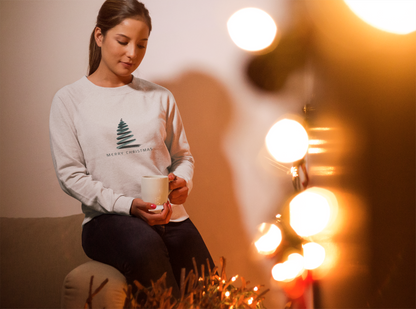 A woman in a white sweater with a Christmas tree design is sitting on a couch, holding a mug and looking down.