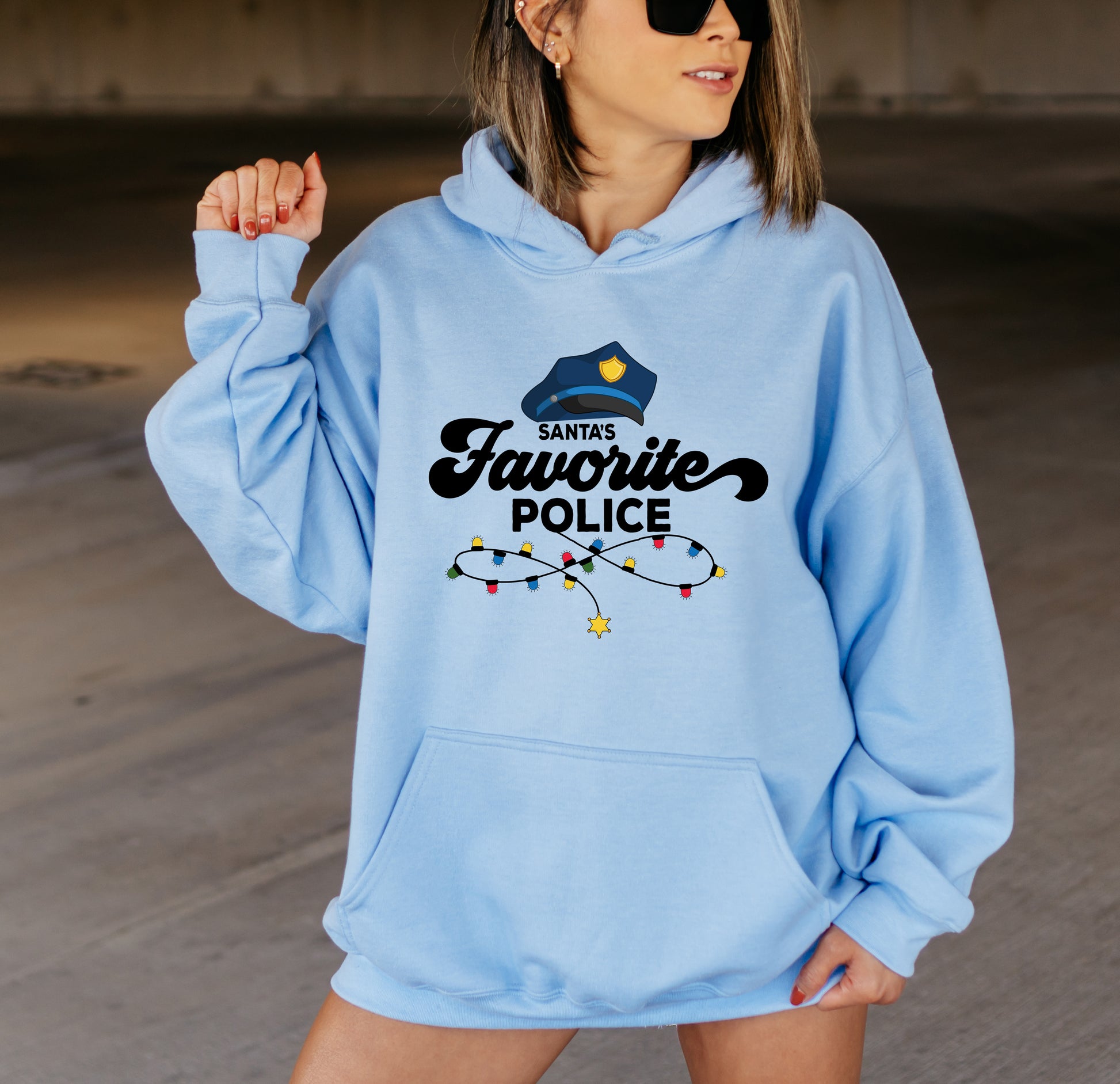 A woman wearing a light blue hoodie with a graphic design that says "Santa's Favorite Police" and a police hat.