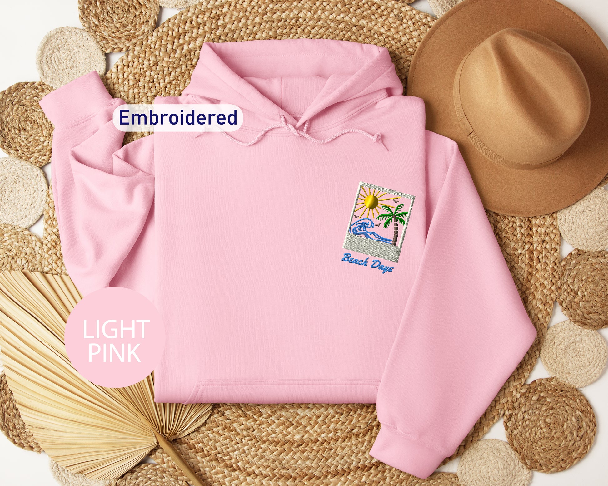 a pink hoodie with a beach-themed embroidered patch, a brown hat, and a straw hat, all arranged on a woven mat.