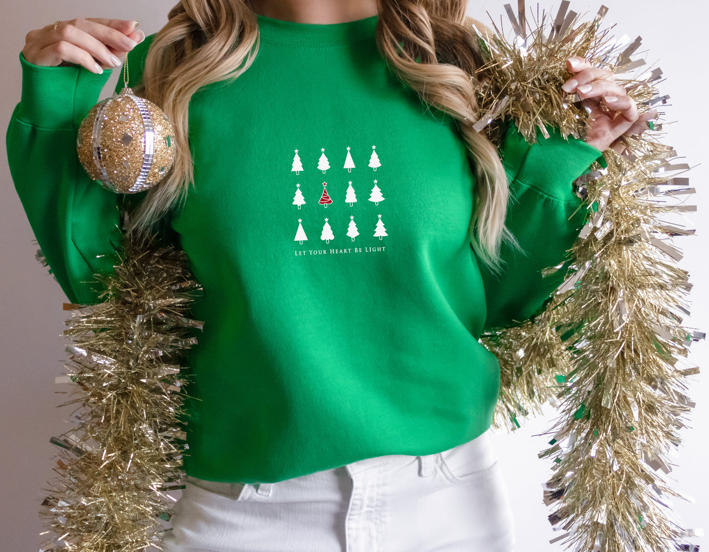 A woman wearing a green sweater with a Christmas tree design and holding a gold ornament and a gold tinsel garland.