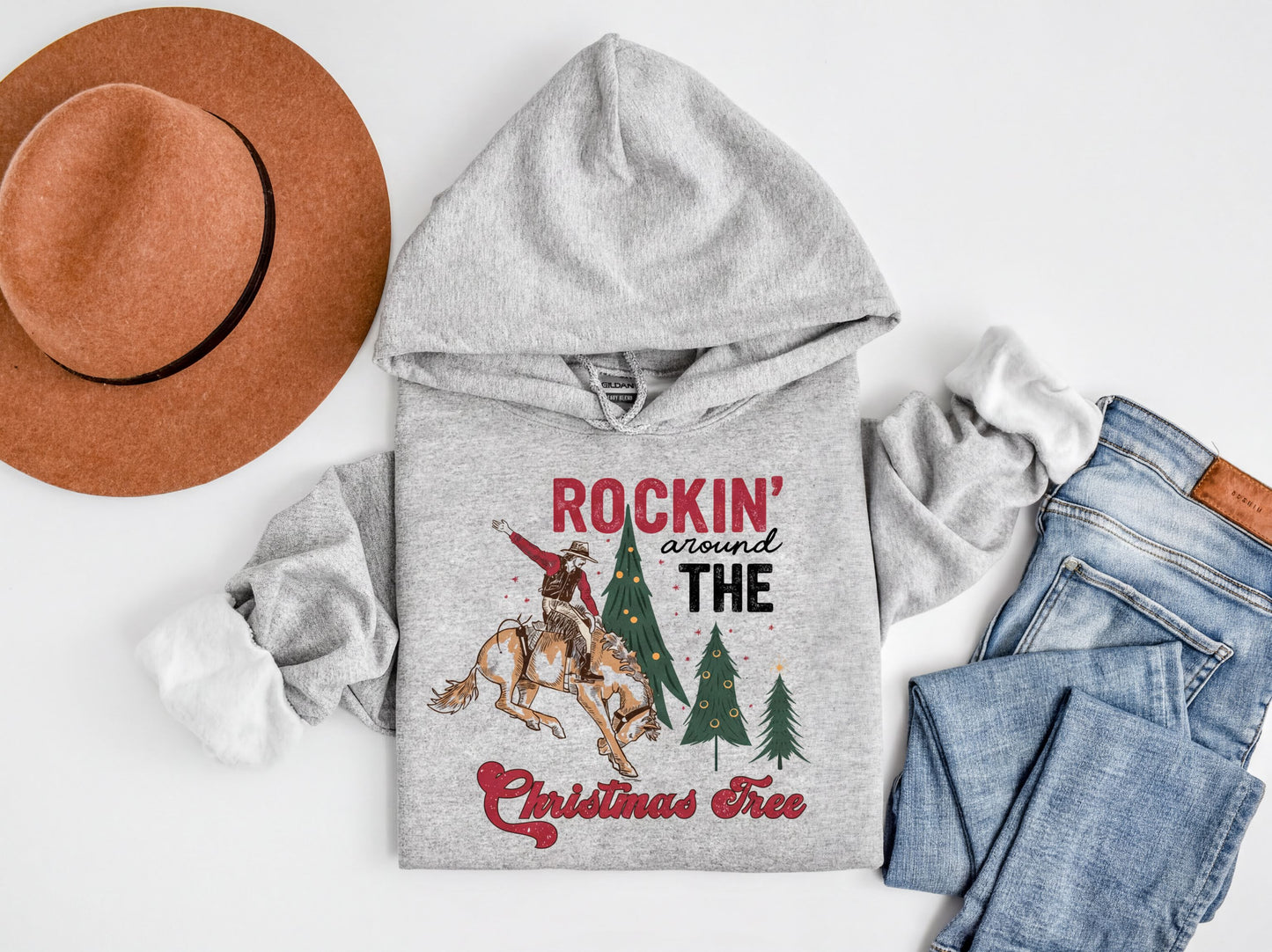 A gray hoodie with a graphic design, a brown hat, a pair of jeans, and a Christmas tree, all laid out on a white background.