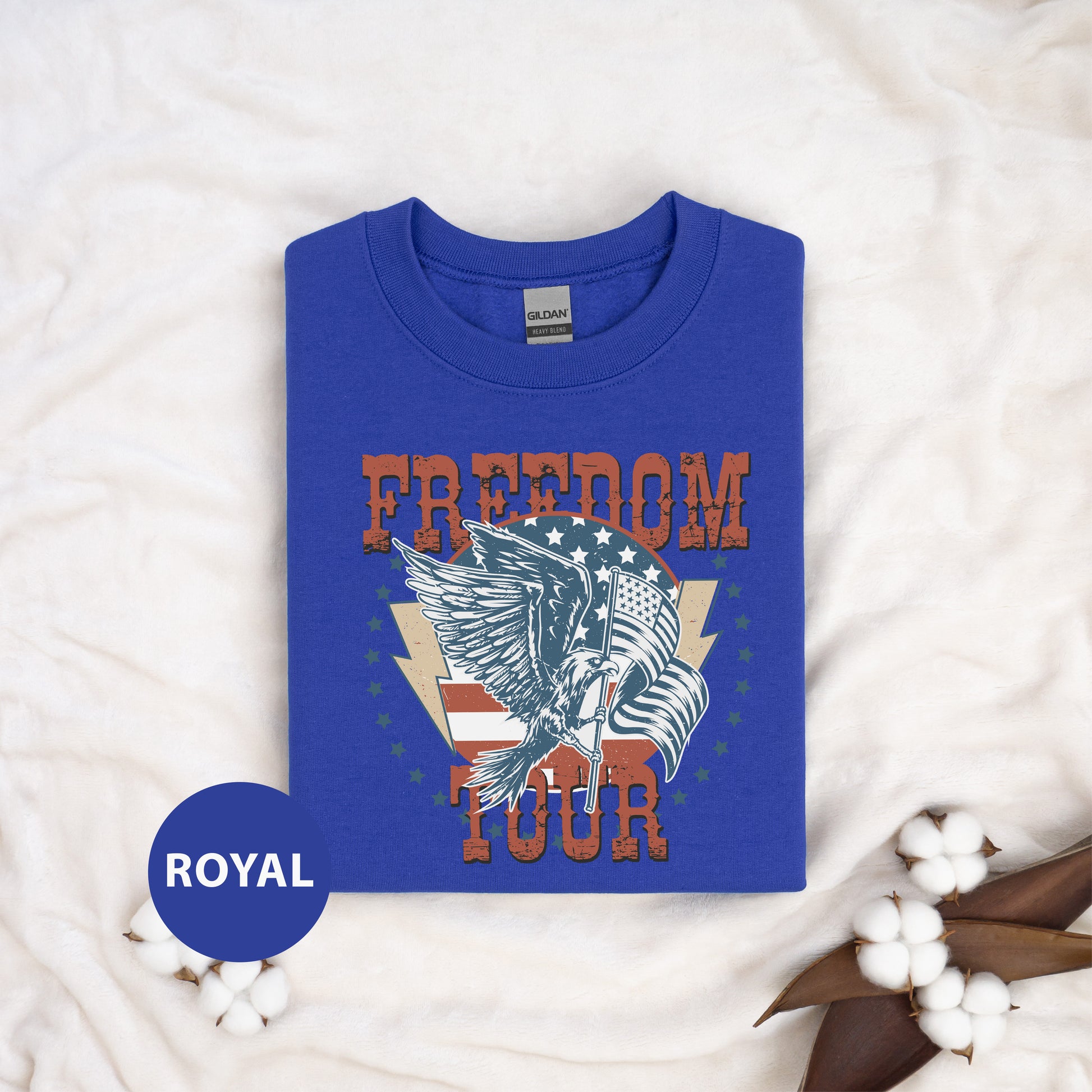 a blue t-shirt with a graphic design of an eagle and the word "FREEDOM" printed on it, along with the word "TOUR" below the eagle. The t-shirt is laid out on a white surface, and there are some cotton balls and leaves in the foreground.