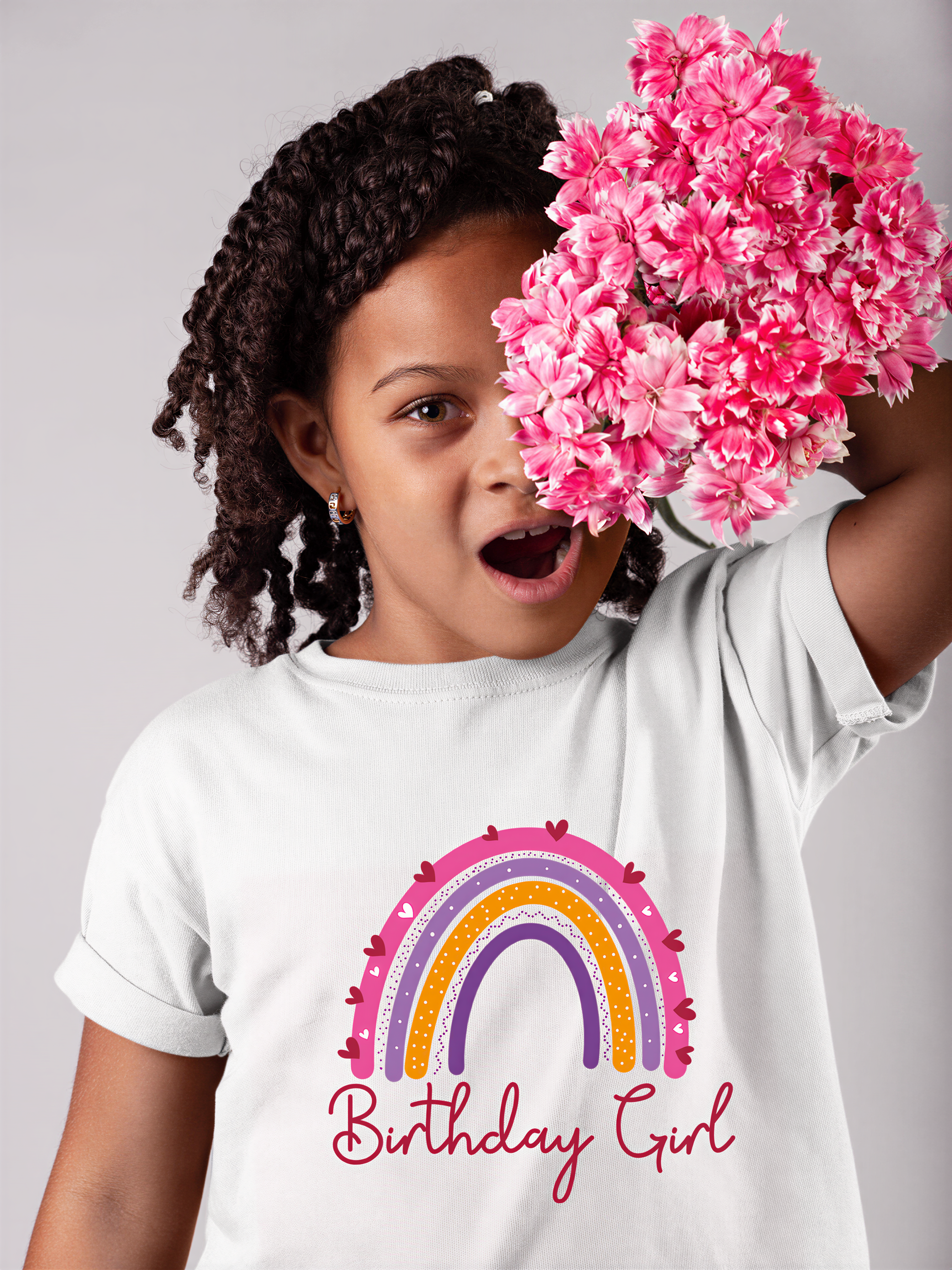 A young girl with curly hair is holding a bouquet of pink flowers up to her face, wearing a white t-shirt with a rainbow graphic and the text "Birthday Girl".