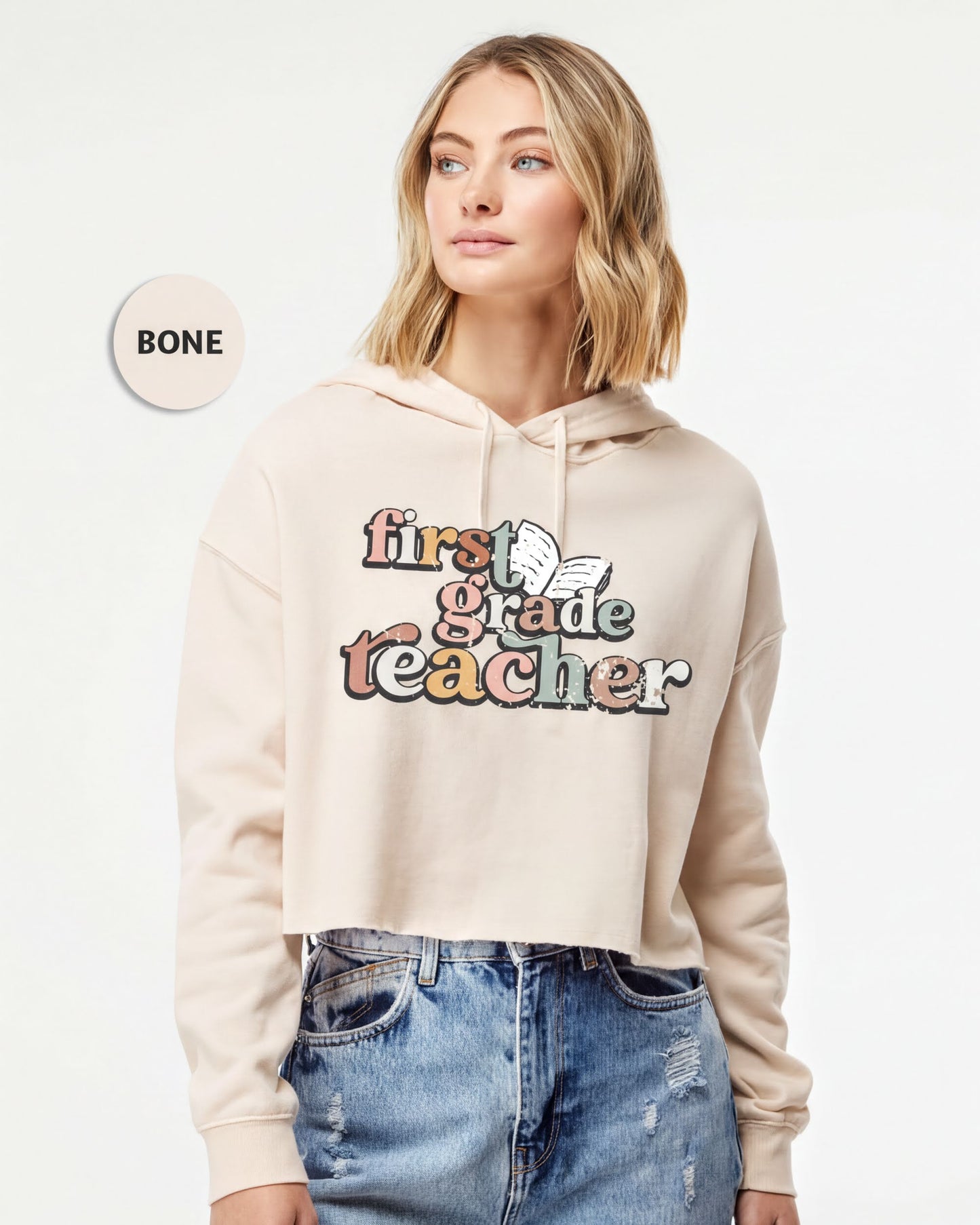 A woman wearing a beige hoodie with the text "first grade teacher" printed on it, standing against a white background.