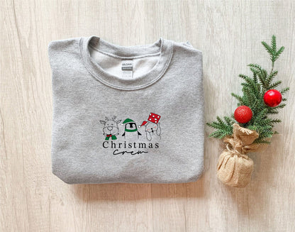 A gray sweatshirt with a Christmas-themed design and text, along with a small Christmas tree and ornaments, are arranged on a wooden surface.