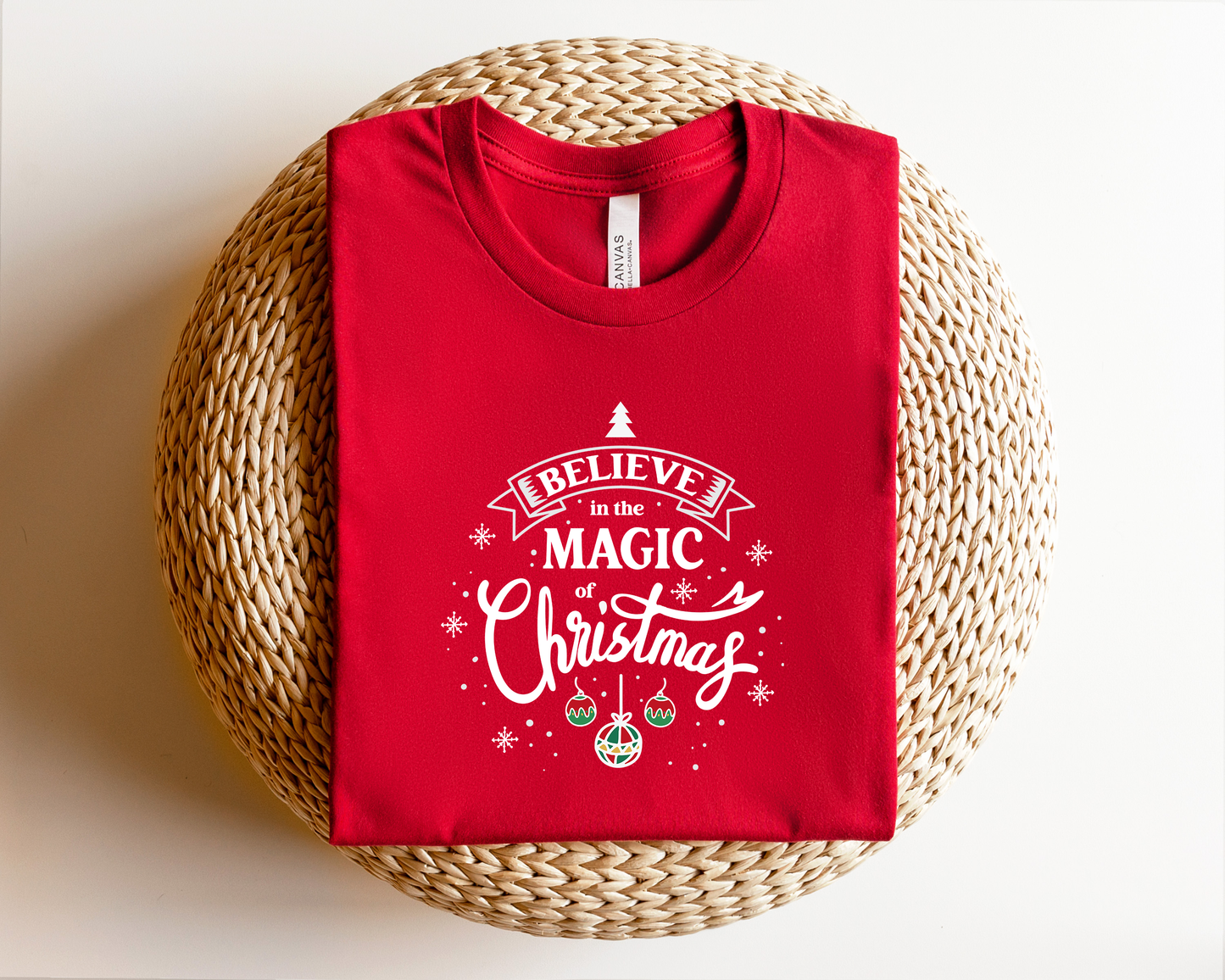 A red t-shirt with a white and red Christmas-themed design and text printed on it, placed on a woven basket.