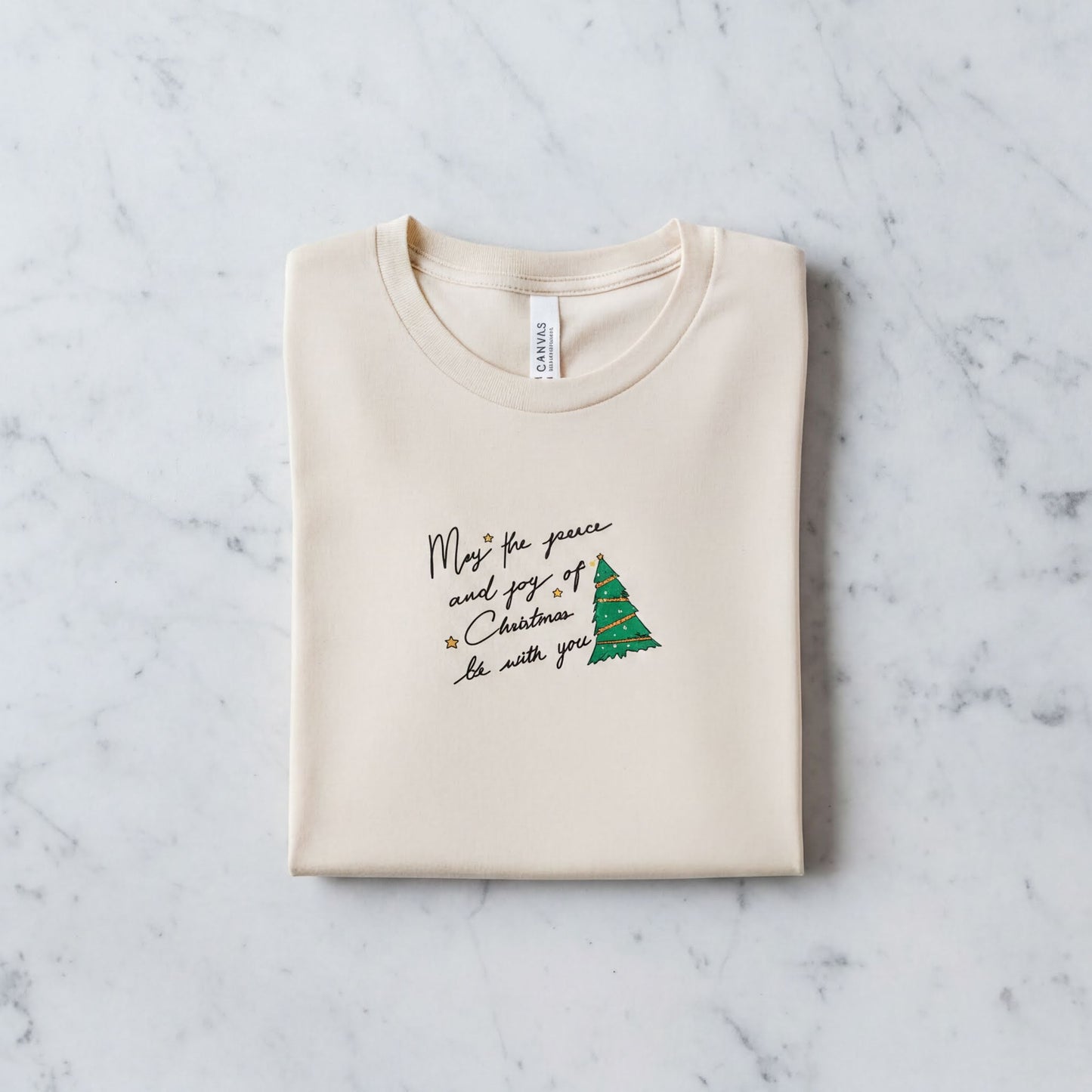 A beige t-shirt with a Christmas-themed design and text printed on it, laid out on a marble surface.