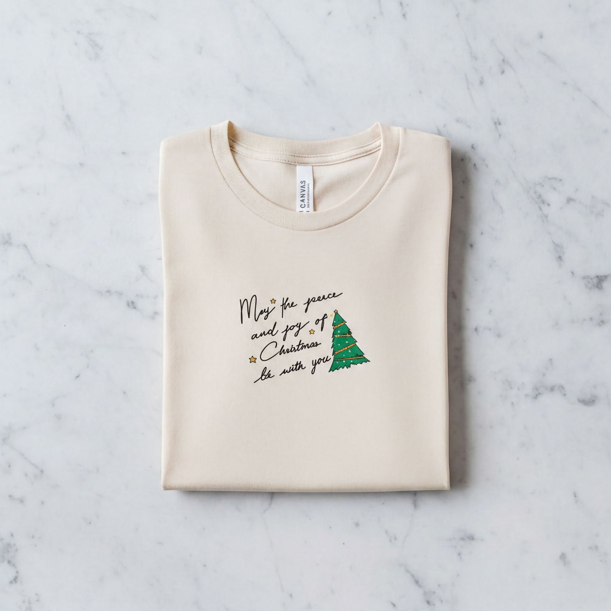 A beige t-shirt with a Christmas-themed design and text printed on it, laid out on a marble surface.