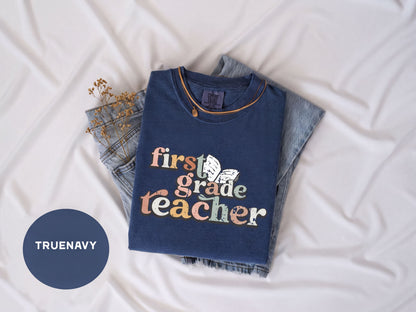 A blue t-shirt with the text "first grade teacher" printed on it, along with a necklace, is laid out on a white surface.