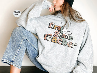 A woman wearing a grey hoodie with the text "first grade teacher" printed on it, sitting against a white background.