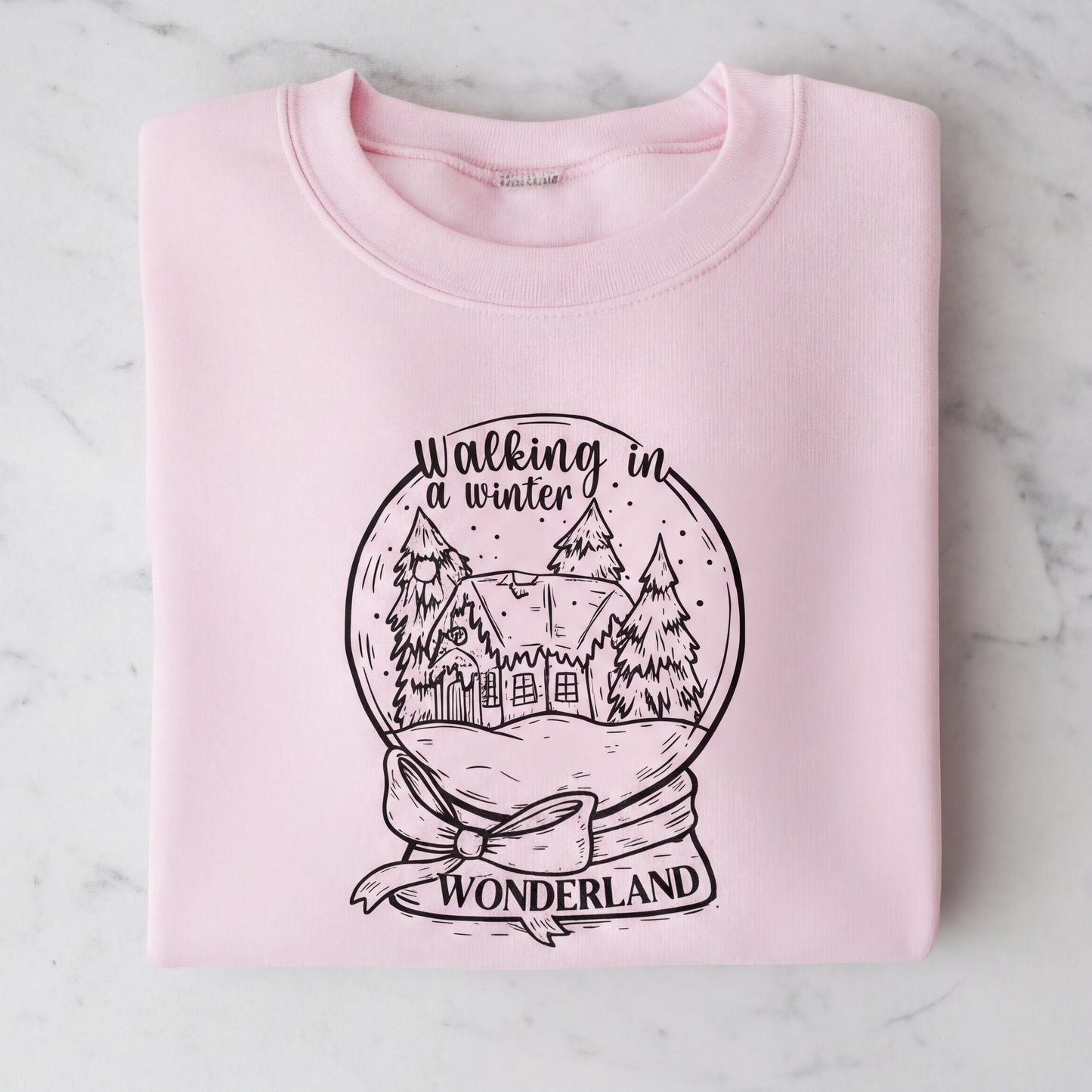A pink sweatshirt with a graphic design depicting a snow globe containing a winter scene with a house and trees. The text on the sweatshirt reads "Walking in winter wonderland".